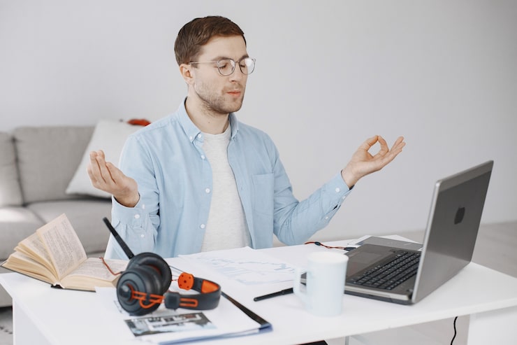 Man practising mindfulness meditation at his desk with a laptop, notebook, and headphones in a home office setting