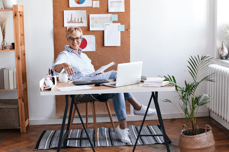 Woman working at a stylish home office desk with a laptop, papers, and corkboard with charts