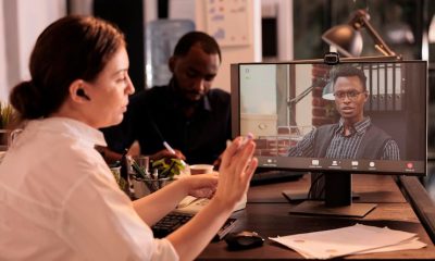 Person on video call in modern office setting, collaborating with colleagues at desk with paperwork and computer