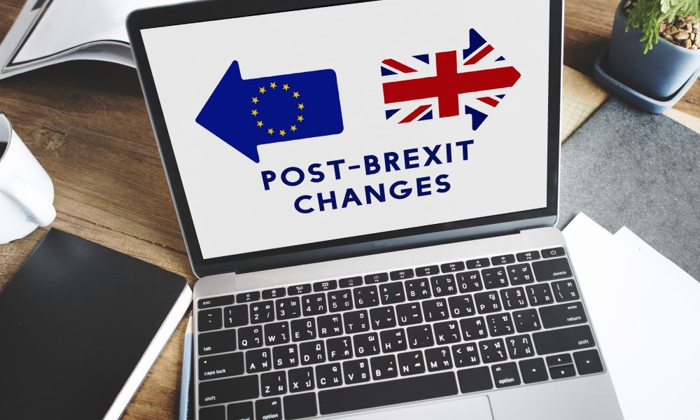 Laptop displaying EU and UK flags with text 'Post-Brexit Changes', beside coffee and notebook on wooden desk