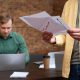 Two men sit at a table, working on laptops and reviewing papers in a collaborative setting