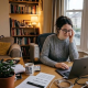 Woman working from home at a laptop, wearing glasses and grey sweater, surrounded by books, plant, and cup of tea
